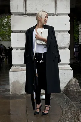 A woman in a black coat, white top, and black pants holds a white bag, standing in front of a stone wall.