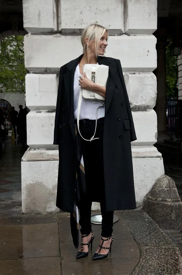 A woman in a black coat, white top, and black pants holds a white bag, standing in front of a stone wall.