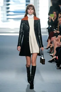 Model walks in Louis Vuitton AW14/15 show wearing a white dress, black leather coat, and knee-high boots.