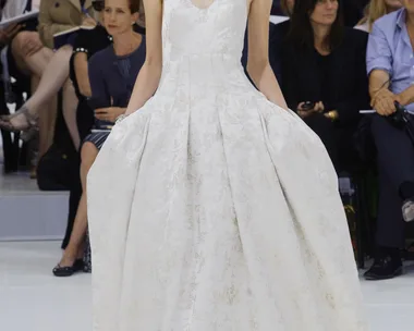 Model in a textured white bridal gown walks runway with audience and floral backdrop.