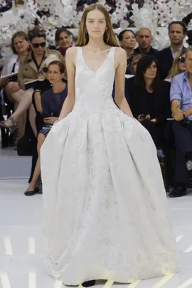 Model in a textured white bridal gown walks runway with audience and floral backdrop.