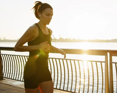 A woman jogging by the river at sunrise, wearing a black fitness outfit and earphones.