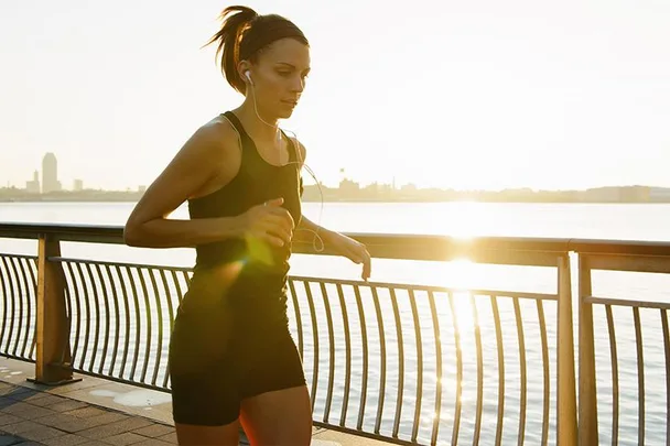 A woman jogging by the river at sunrise, wearing a black fitness outfit and earphones.