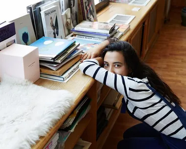 A woman in a striped shirt leans on a wooden shelf filled with books and magazines, looking at the camera.