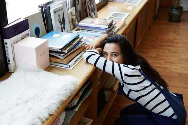 A woman in a striped shirt leans on a wooden shelf filled with books and magazines, looking at the camera.