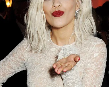 Blonde woman in lace dress blows a kiss, wearing bold red lipstick and matching nails, with wavy hair and earrings.