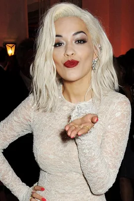 Blonde woman in lace dress blows a kiss, wearing bold red lipstick and matching nails, with wavy hair and earrings.