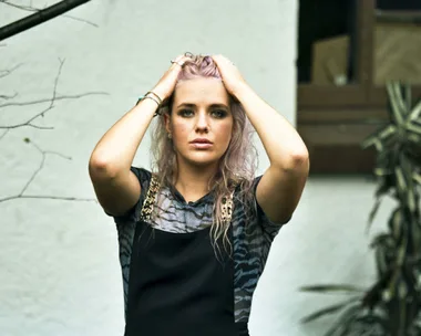 Woman with platinum blonde hair holds her head with both hands, standing outdoors with a neutral expression.