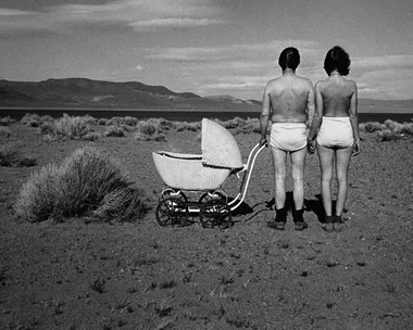 Two people in underwear holding hands, standing next to an empty baby carriage in a desolate, dry landscape.