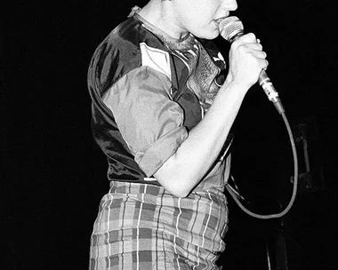 A person in a plaid skirt and short-sleeved shirt sings into a microphone on a dark stage.