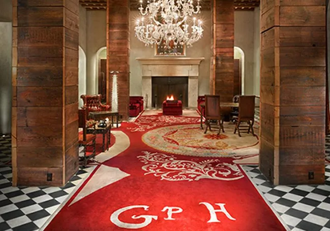 Luxurious hotel lobby with red carpet, chandelier, wooden columns, and a lit fireplace. Text "GPH" visible on carpet.