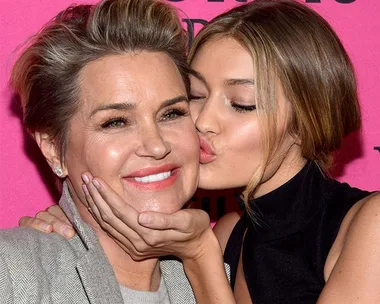 A woman kisses another woman on the cheek, both smiling, with a pink background.