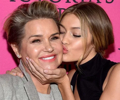 A woman kisses another woman on the cheek, both smiling, with a pink background.