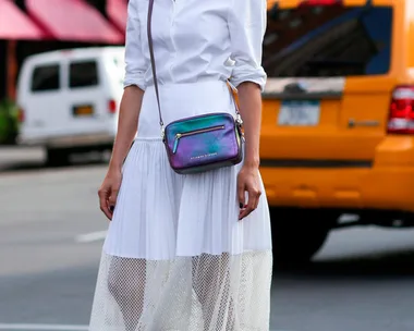 Woman in white dress and red sunglasses, with a purple crossbody bag, stands in front of a yellow taxi in a city street.