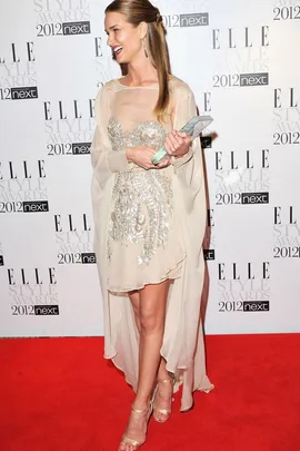 Person in a sequined beige dress holding an award, smiling on a red carpet at the 2012 Elle Style Awards.