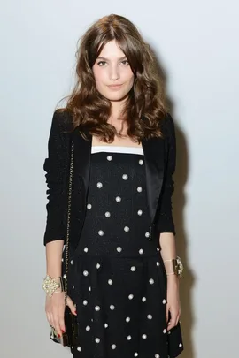 A woman with wavy brown hair in a black dress with pearl accents, standing against a light background.