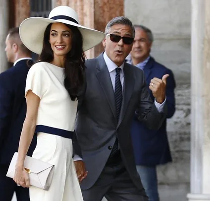 A couple in formal attire, with the man gesturing happily near a building, surrounded by people.