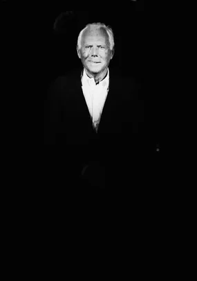 Black-and-white image of an elderly man in a white shirt and black jacket, highlighted in spotlight against a dark background.