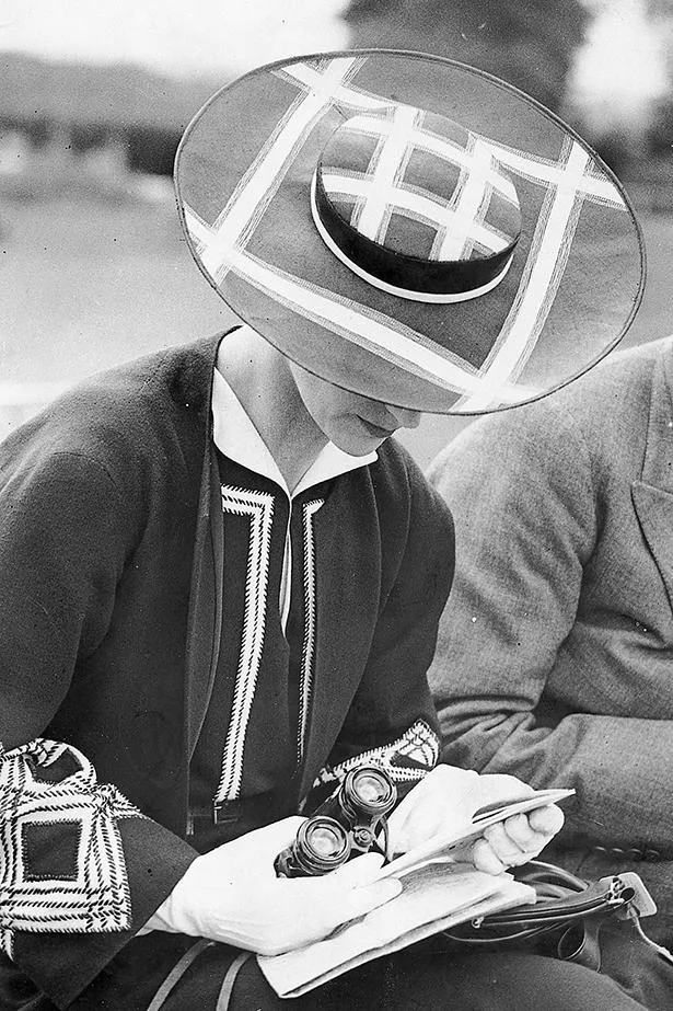 A woman in vintage attire and a stylish hat holds binoculars and a race program.