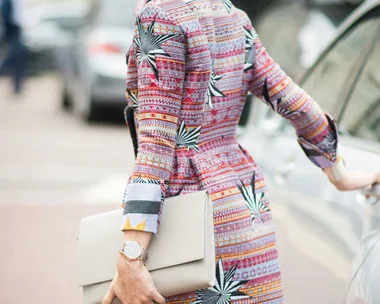 Woman in a colorful patterned coat, holding a white clutch, with hair in a bun and wearing sunglasses, standing by a car.