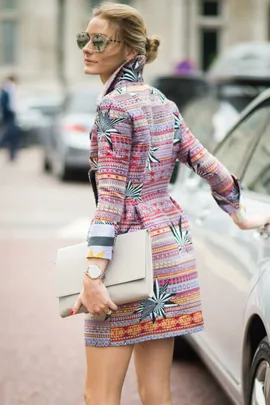 Woman in a colorful patterned coat, holding a white clutch, with hair in a bun and wearing sunglasses, standing by a car.