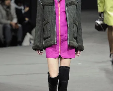 Model on runway in a pink and black outfit with a puffer jacket, knee-high boots, and slicked-back hair.