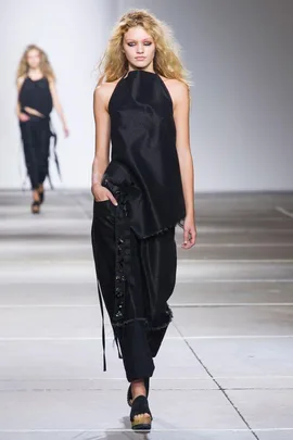 Model walking on a runway wearing a black asymmetrical top and pants, with long wavy hair, in a Marques Almeida show.