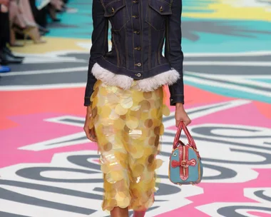 Model walks runway in denim jacket, yellow sequin skirt, and colorful bag at Burberry Prorsum SS15 show.