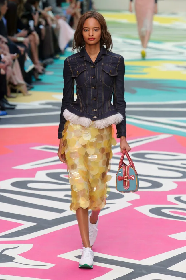 Model walks runway in denim jacket, yellow sequin skirt, and colorful bag at Burberry Prorsum SS15 show.