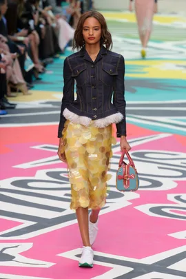 Model walks runway in denim jacket, yellow sequin skirt, and colorful bag at Burberry Prorsum SS15 show.