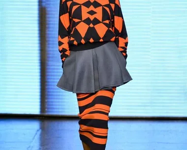 Model in a geometric orange and black sweater, gray skirt, and striped skirt walks a runway at DKNY Spring/Summer 2015.