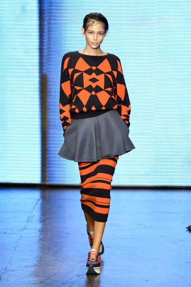 Model in a geometric orange and black sweater, gray skirt, and striped skirt walks a runway at DKNY Spring/Summer 2015.