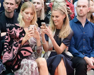 Two women sit together in a crowd, looking at their smartphones and smiling; others around appear serious.