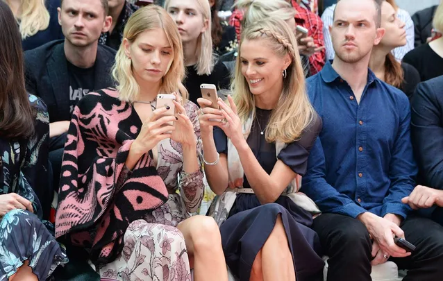 Two women sit together in a crowd, looking at their smartphones and smiling; others around appear serious.