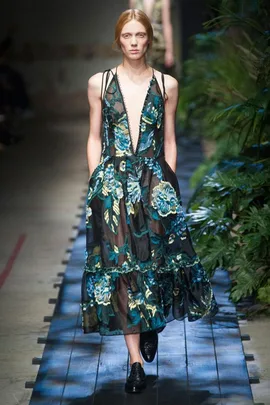 Model walks runway in sheer floral Erdem dress with deep V-neck and black shoes, surrounded by lush greenery.