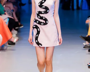 Model walks runway in a pale pink dress with black sequin wave design and yellow shoes at a fashion show.