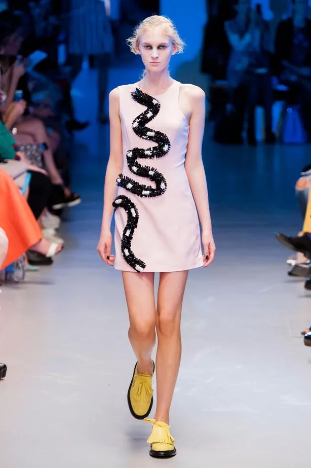 Model walks runway in a pale pink dress with black sequin wave design and yellow shoes at a fashion show.