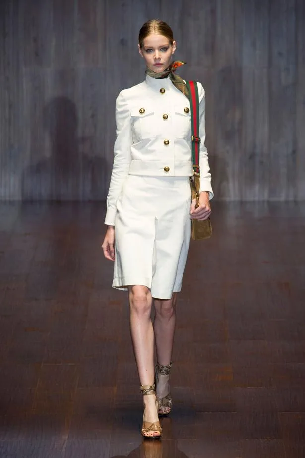 Model in white outfit with gold buttons, high-neck top, pencil skirt, sandals, and a crossbody. Gucci SS15 fashion show.