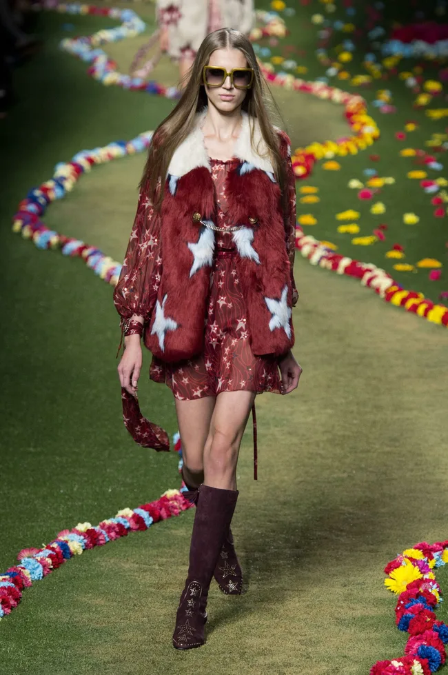 Model walking runway in a red star-patterned dress, fur vest, and sunglasses; surrounded by colorful flower petals.