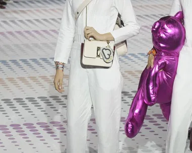 Model wearing a white outfit with a crossbody bag and holding a pink inflatable bear on a dotted runway.