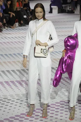 Model wearing a white outfit with a crossbody bag and holding a pink inflatable bear on a dotted runway.