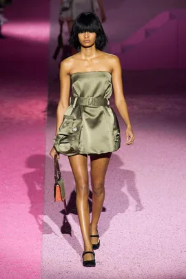 Model on a pink runway wearing a strapless, olive mini dress with pockets, holding a brown handbag.