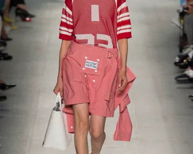 Model walks runway in red sports jersey and pink shorts with white tote bag, wearing white strappy sandals.