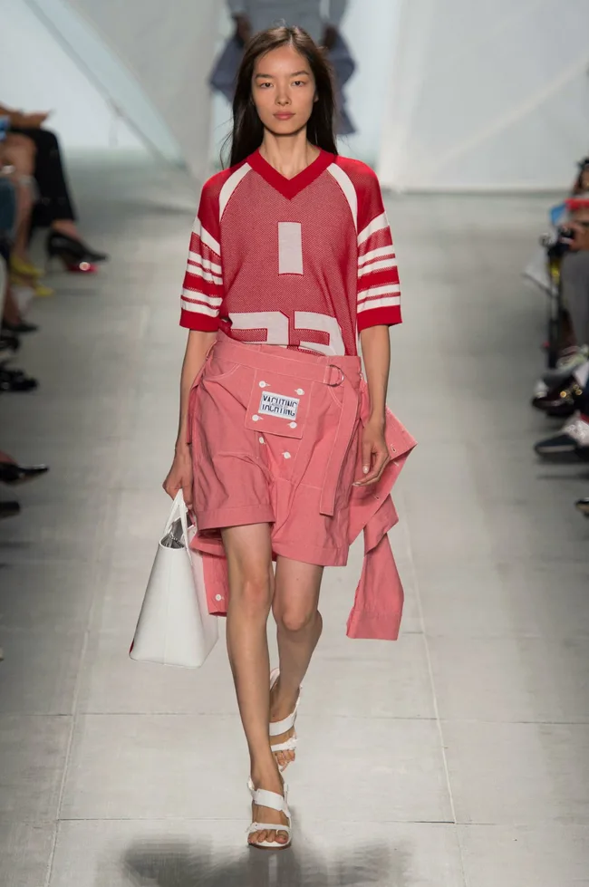 Model walks runway in red sports jersey and pink shorts with white tote bag, wearing white strappy sandals.