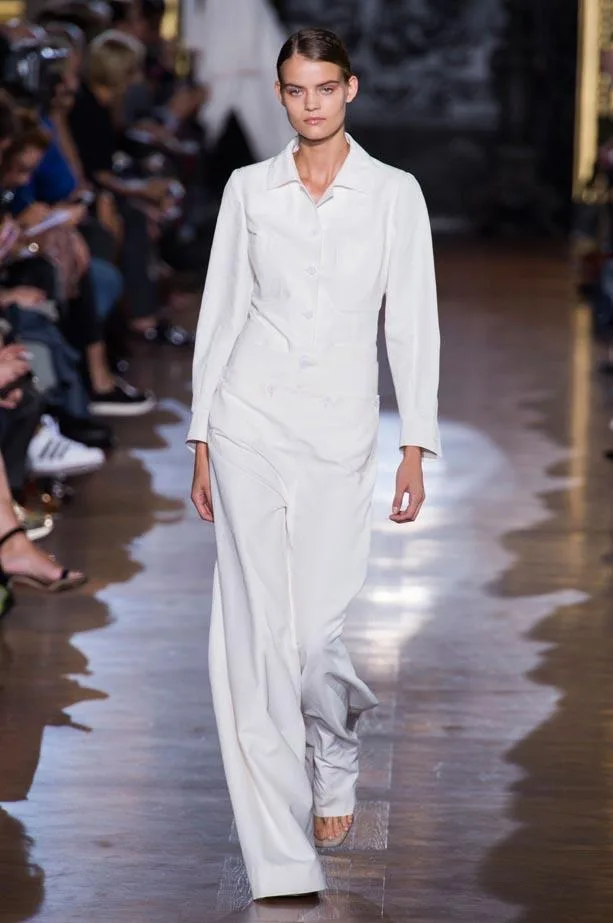 Model walking in Stella McCartney Spring/Summer 2015 show wearing a white tailored suit on a wooden runway.