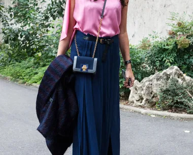 Woman in pink top, blue skirt, carrying a navy purse, holding a coat, walking on a street lined with greenery.