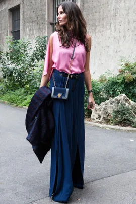 Woman in pink top, blue skirt, carrying a navy purse, holding a coat, walking on a street lined with greenery.