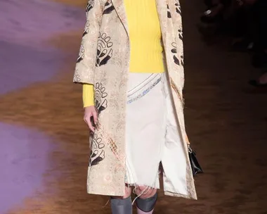 Model walks runway in Prada's SS15 show wearing a printed coat, yellow sweater, knee-length skirt, and styled black shoes.