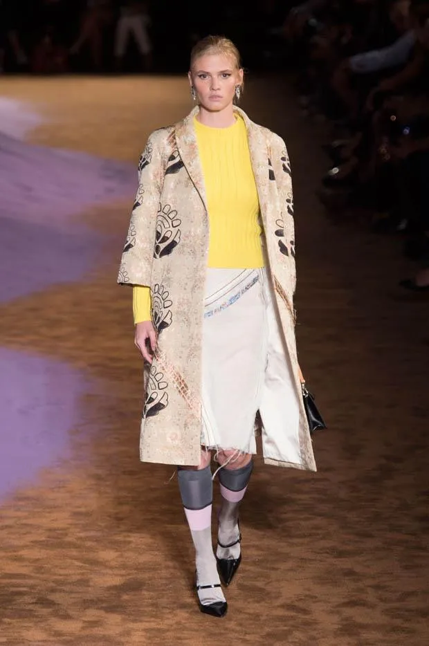 Model walks runway in Prada's SS15 show wearing a printed coat, yellow sweater, knee-length skirt, and styled black shoes.