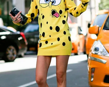 Person wearing a yellow sweater dress with a cartoon face design, posing on a city street with taxis in the background.